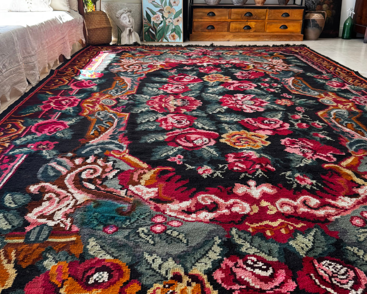 A large rectangular area rug with an ornate floral and scroll pattern in red, pink, orange, teal, and cream on a dark background, shown in a styled living room setting.