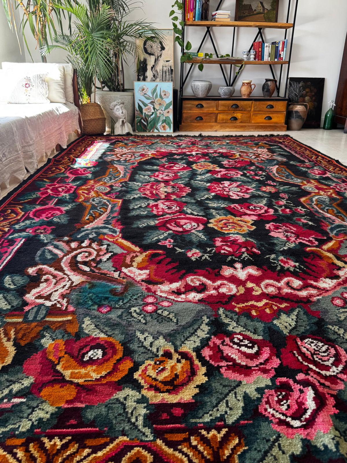 A large rectangular area rug with an ornate floral and scroll pattern in red, pink, orange, teal, and cream on a dark background, shown in a styled living room setting.