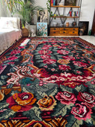 A large rectangular area rug with an ornate floral and scroll pattern in red, pink, orange, teal, and cream on a dark background, shown in a styled living room setting.